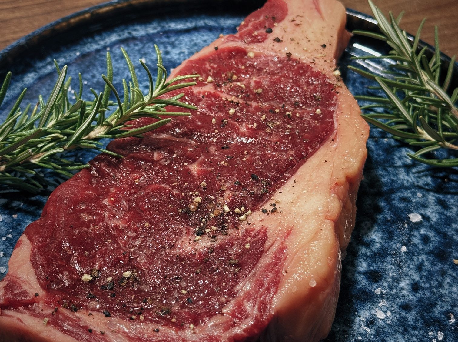 Raw rib eye steak seasoned with salt and pepper, with rosemary sprigs on a blue ceramic plate